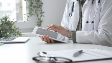 Beyaz laboratuvar önlüğü ve steteskop giyen profesyonel kadın doktor, klinik ortamdaki hasta kayıtlarını incelerken dijital tablet üzerinde çalışıyor. İlaç ve sağlık hizmetleri kavramı