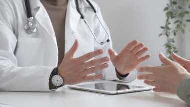 Laboratuvar önlüğü ve steteskop giymiş bir erkek doktor modern tıp ofisinde tablet kullanan hastaya teşhis koymayı açıklarken el kol hareketi yapıyor. İlaç ve sağlık hizmetleri kavramı.