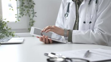 Beyaz laboratuvar önlüğü ve steteskop giyen profesyonel kadın doktor, klinik ortamdaki hasta kayıtlarını incelerken dijital tablet üzerinde çalışıyor. İlaç ve sağlık hizmetleri kavramı