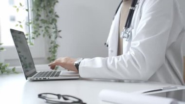 Laptop üzerinde çalışan, modern, iyi aydınlatılmış sağlık ortamında beyaz önlük ve steteskop giyen, klinik uzmanlığı dijital teknolojiyle harmanlayan bir tıp uzmanı. İlaç ve sağlık hizmetleri