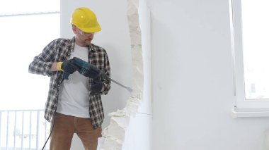 Bej kareli gömlek, sarı başlık ve koruyucu eldiven giyen inşaat işçisi, dönen çekiç matkabıyla beyaz duvarı yıkıyor ve orta uzunlukta toz üretiyor. Yenileme konsepti