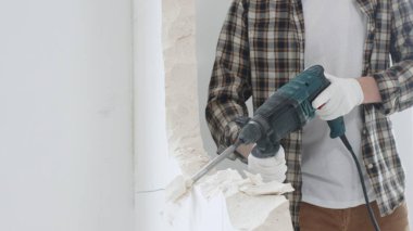 Tanınmayan bej kareli gömlek ve koruyucu eldivenler giyen erkek inşaat işçisinin yakın çekiç matkabıyla beyaz duvarı yıkıyor, toz üretiyor, yakın görüş sağlıyor. Yenileme