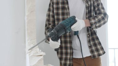 Tanınmayan bej kareli gömlek ve koruyucu eldivenler giyen erkek inşaat işçisinin yakın çekiç matkabıyla beyaz duvarı yıkıyor, toz üretiyor, yakın görüş sağlıyor. Yenileme.