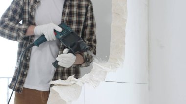 Tanınmayan bej kareli gömlek ve koruyucu eldivenler giyen erkek inşaat işçisinin yakın çekiç matkabıyla beyaz duvarı yıkıyor, toz üretiyor, yakın görüş sağlıyor. Yenileme.
