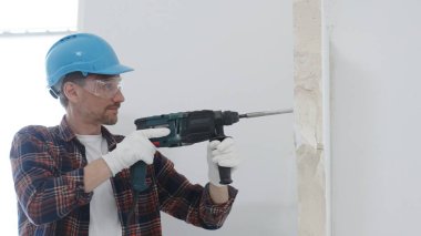 Kırmızı kareli gömlek, mavi koruyucu kask ve eldiven giyen erkek inşaat işçisi, döner çekiç matkabıyla beyaz duvarı yıkıyor, toz ve portre görüntüsü yaratıyor. Yenileme konsepti.