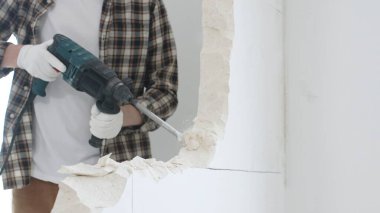 Tanınmayan bej kareli gömlek ve koruyucu eldivenler giyen erkek inşaat işçisinin yakın çekiç matkabıyla beyaz duvarı yıkıyor, toz üretiyor, yakın görüş sağlıyor. Yenileme