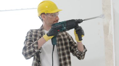 Bej kareli gömlek, sarı başlık ve koruyucu eldiven giyen bir inşaat işçisinin portresi, döner çekiç matkabıyla beyaz duvarı yıkıyor ve toz oluşturuyor. Yenileme konsepti.