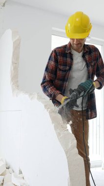 Kırmızı kareli gömlek, sarı başlık ve koruyucu eldiven giyen erkek inşaat işçisi, dönen çekiç matkabıyla beyaz duvarı yıkıyor ve orta uzunlukta toz üretiyor. Yenileme projesi.