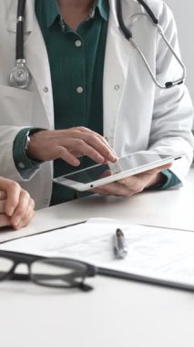 Yeşil gömlekli bir erkek doktor ve modern tıp ofisinde tablet kullanan hastaya teşhisi açıklarken steteskop hareketi yapıyor. İlaç ve sağlık hizmetleri kavramı.