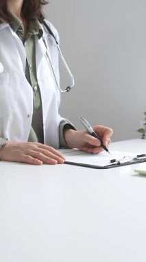 Laboratuvar önlüğü ve steteskop giymiş kadın doktor, tıp bürosunun masasındaki panoya profesyonelliği vurgulayan dizüstü bilgisayar ve gözlüklerle notlar yazıyor. İlaç ve sağlık hizmetleri kavramı