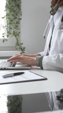 Stetoskop ve laboratuvar önlüğü giyen doktor özenle bir dizüstü bilgisayarda yazıyor ve modern bir tıp ofisinde verimli bir sağlık hizmeti yönetimi sergiliyor. İlaç ve sağlık hizmetleri kavramı