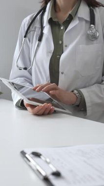 Beyaz laboratuvar önlüğü ve steteskop giyen profesyonel kadın doktor, klinik ortamdaki hasta kayıtlarını incelerken dijital tablet üzerinde çalışıyor. İlaç ve sağlık hizmetleri kavramı