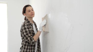 Kareli gömlek giyen kadın inşaat işçisi, beyaz duvar yüzeyini düzleştiriyor, profesyonel spatula tekniği ile macunu uyguluyor, iç dekorasyon konusunda yetenekli..