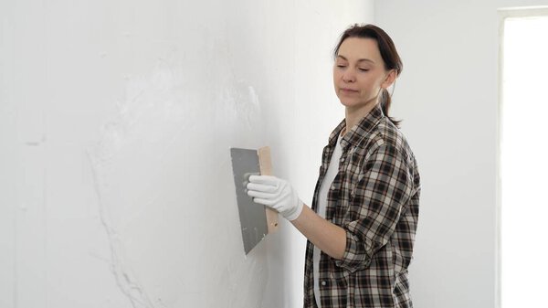 Female construction professional wearing checkered shirt is smoothing white wall surface, applying putty with professional spatula technique, showcasing skilled interior renovation work.
