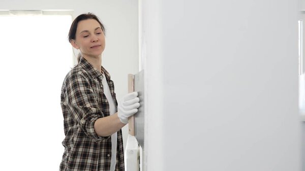 Female construction professional wearing checkered shirt is smoothing white wall surface, applying putty with professional spatula technique, showcasing skilled interior renovation work.