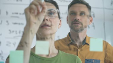 Businesswoman wearing glasses, writing on a glass board and organizing sticky notes, collaborates with adult businessman observing her strategy planning in a bright office.