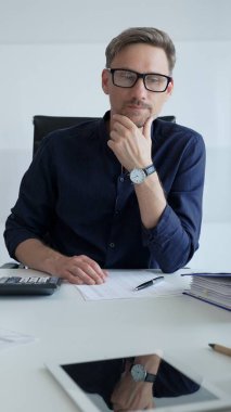 Businessman sitting at a sleek desk, working on a laptop and thinking in a bright, modern office filled with natural light. Business people concept.
