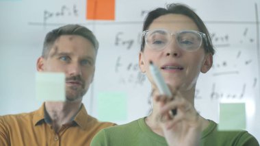 Businesswoman holding a marker while explaining strategy to a colleague, using a glass board filled with sticky notes during an engaging work meeting in a corporate office setting