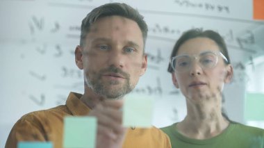 Two business colleagues are actively engaged in a brainstorming session, utilizing sticky notes on a glass board to organize their thoughts and develop a strategic plan for their project