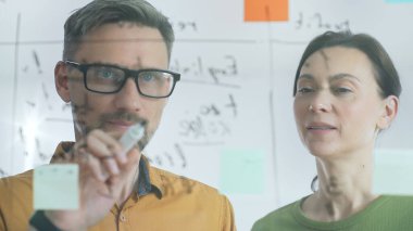 İki iş arkadaşı, bir beyin fırtınası sırasında şeffaf bir tahtayla ilgili notları analiz ediyor. Anlaşmaları için yenilikçi çözümler ve stratejiler geliştirmek için birlikte çalışıyorlar..