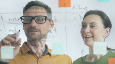 İki iş arkadaşı, bir beyin fırtınası sırasında şeffaf bir tahtayla ilgili notları analiz ediyor. Anlaşmaları için yenilikçi çözümler ve stratejiler geliştirmek için birlikte çalışıyorlar..