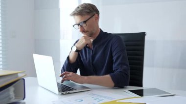 Businessman sitting at a sleek desk, working on a laptop and thinking in a bright, modern office filled with natural light. Business people concept.