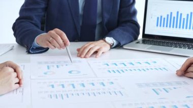 İş arkadaşları, bir toplantı sırasında mali raporları ve kağıt tablolardaki kurumsal verileri analiz ederek, pazar stratejisi ve proje sonuçlarını tartışarak, bir ofis ortamında