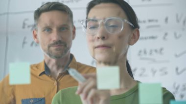 Businesswoman wearing glasses, writing on a glass board and organizing sticky notes, collaborates with adult businessman observing her strategy planning in a bright office.