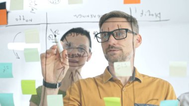 İki işadamı, beyin fırtınası sırasında renkli yapışkan notlarla kaplı cam bir tahtaya yazarak şirketleri için yeni fikirler ve stratejiler geliştiriyor.