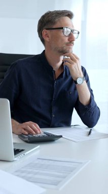 Businessman sitting at a sleek desk, working on a laptop and thinking in a bright, modern office filled with natural light. Business people concept.