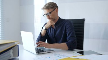 Professional businessman sitting at a sleek desk, working on a laptop and thinking about goals in modern office filled with natural light. Business people concept.