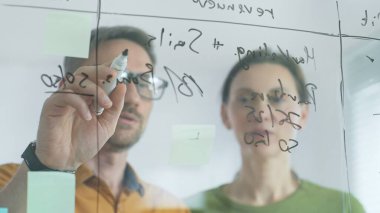 Business people brainstorming near glass board, using sticky notes for strategic planning, writing calculations, sharing innovative ideas during collaborative work session. close up.