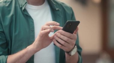 Close-up view of attractive young caucasian mans hands using smartphone online checking social media making internet payments. Focused modern adult male typing texting scrolling on phone outside.