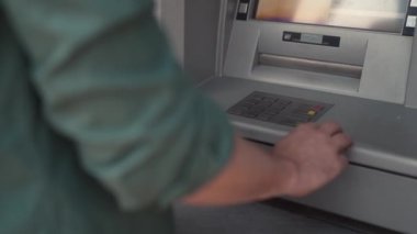 Close-up view of young adult caucasian man using ATM to check balance of credit debit card on street. Male client of bank making withdrawal, payments, transactions. Finances concept.