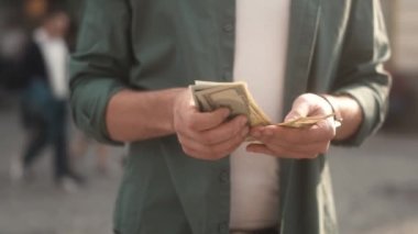 Close-up view of hands of successful caucasian man counting dollar bills standing on crowded street with people. Young adult stylish male holding salary income after withdrawal. Finance concept.