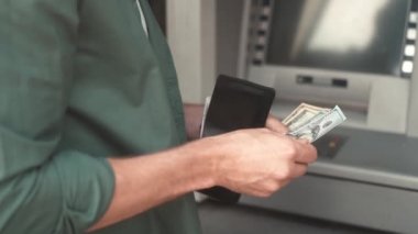 Close-up view of hands of adult young man receiving money from ATM. Modern male withdrawing dollar bills counting putting in wallet. Making currency financial operations and transactions.