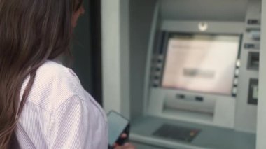Back view of young caucasian female using ATM terminal withdrawing money on street. Adult woman making bank transactions cashing out holding receipt of bank operation. Finance concept.
