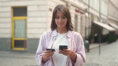 Portrait of attractive amazed caucasian woman using typing on smartphone doing shopping online. Beautiful happy female holding phone and debit card making successful payment smiling outside.