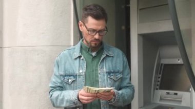 Portrait of young successful businessman counting dollar bills withdrawn from bank card using ATM terminal. Attractive adult caucasian male holding money after making financial operations.