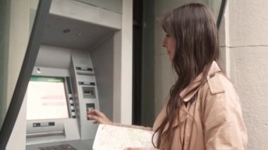 Side-view portrait of young pretty woman tourist inserting bank card making financial operations using ATM terminal on street. Attractive beautiful caucasian female making transactions,