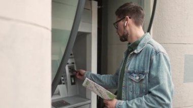 Side-view portrait of young attractive modern man inserting bank card in ATM terminal entering password making financial payments outside. Beautiful male tourist holding man wearing earphones.