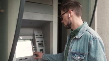 Side-view portrait of young adult caucasian tourist using ATM to withdraw money from bank card check balance. Attractive modern man standing outside listening music in earphones holding map.