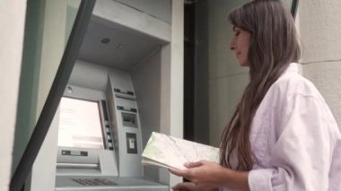 Low angle side view portrait of focused caucasian female tourist with map using ATM to make bank transactions outside. Stylish woman wearing coat making bank operations entering pincode.