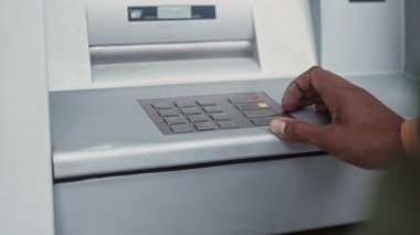 Close-up view of young African-American mans hands entering pincode of bank card using ATM terminal to make financial operations. Adult African male making transactions outdoors.