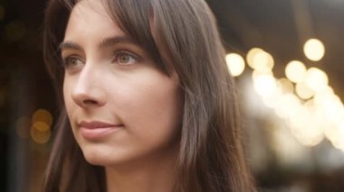 Close-up portrait of young thoughtful cute woman with brown eyes looking. Face of attractive pretty adult caucasian female standing in cozy cafe terrace with blurred fairy lights on background.