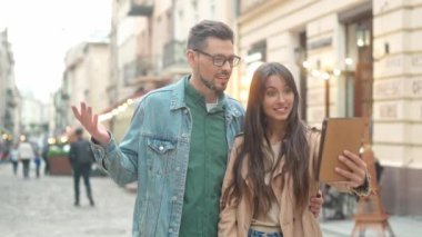 Portrait of two attractive young adult caucasian people talking online video calling with tablet standing in city center. Handsome man and beautiful woman holding device communicating.