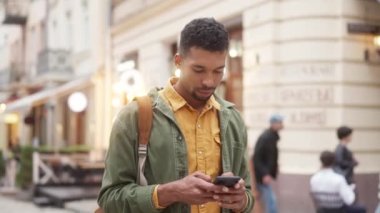 Portrait of handsome focused young African man standing in city center texting with smartphone wearing stylish clothes. Attractive African American male tourist communicating online.