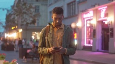 Portrait of focused relaxed young African American male tourist communicating online texting with phone standing in city center with beautiful neon signs on background. Evening concept.