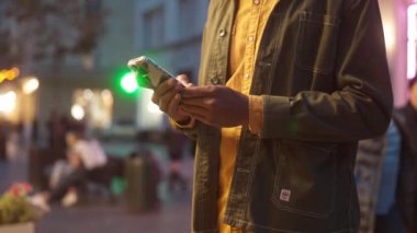 Low angle view portrait of focused African American man using smartphone checking social media sending messages online. Handsome busy male tourist standing on street with neon signs.