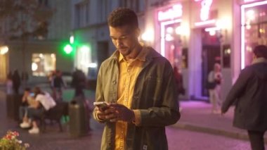 Portrait of handsome cheerful man texting friends online with smartphone looking at camera smiling. Beautiful happy African male tourist standing on street with neon signs in evening.